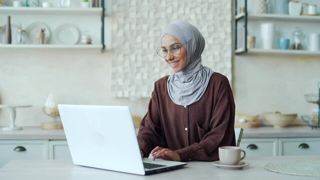 Smiling Muslim woman in hijab enjoying cup of coffee or tea and continue her work typing on laptop in light room Attractive female doing job browsing web at kitchen Online studying or work from home