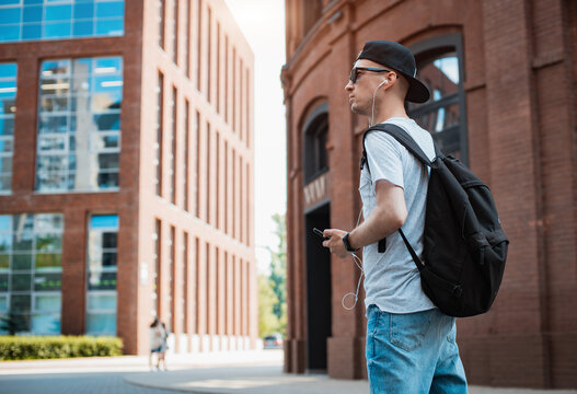 young man is a fashionable stylish guy in sunglasses and a black cap walking around the city holding a smartphone in his hand and listening to music headphones. T