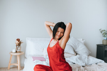 Young brunette hispanic woman in red pyjamas stretching sitting on bed at home on Sunday morning....