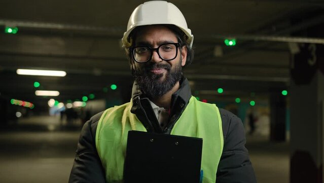 Indian contractor mechanic guy in helmet and glasses smile posing with clipboard folder. Arabian bearded man in eyeglasses worker male specialist manufacture work. Engineer in hard hat safety uniform
