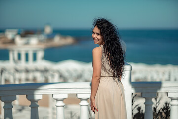 Fototapeta na wymiar Sea woman rest. A woman with long curly hair in a beige dress stands with her back and looks at the sea and the coast from a balcony with balusters. Tourist trip to the sea.