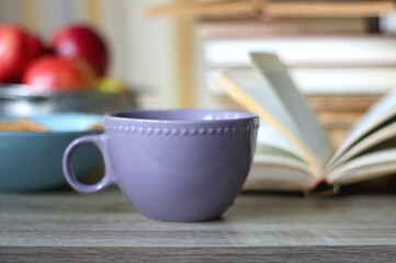 Stack of books, e-reader, reading glasses, bowl of biscuits, apples and cup of tea on the table. Bookshelf in the background. Selective focus.