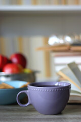Stack of books, e-reader, reading glasses, bowl of biscuits, apples and cup of tea on the table. Bookshelf in the background. Selective focus.