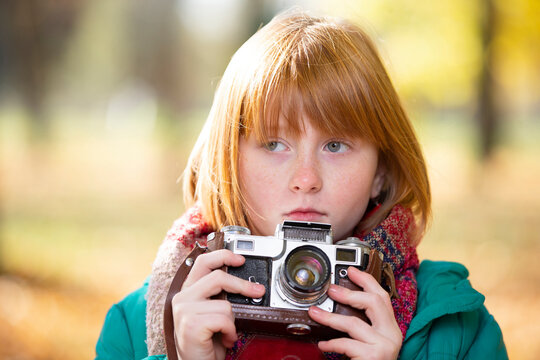 Little red-haired girl with a retro camera in the autumn park. Child photographer.