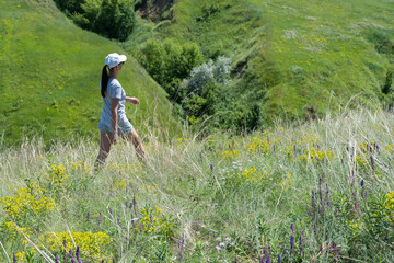 Woman goes among flowers and looking on ravine covered with greenery. Young female traveler step...
