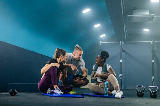 Fitness gym instructor showing workout plan on tablet to two fit woman in sportswear with towel. They are sitting on the gym floor, resting after workout.
