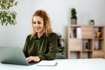 A smiling female employee working over the laptop, sitting at the home office.