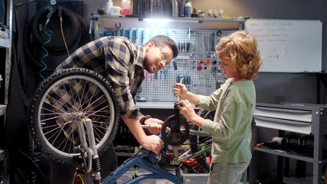 Beautiful caring skillful dad teaching cute little boy to fix bike in authentic workshop with instruments. Attractive caucasian son using tools to repair bicycle with young adult father. Parenting.