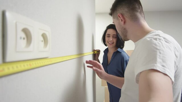 Handheld video of a couple taking measurements on a wall. A young couple is discussing apartment renovation. Happy man and woman in love using measuring equipment measure the wall.