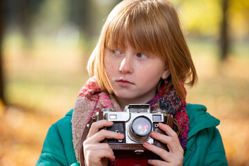 Little red-haired girl with a retro camera in the autumn park. Child photographer.