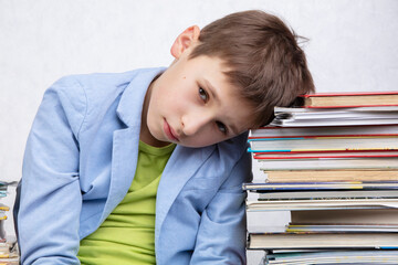 A tired school-age boy sits between stacks of books. Sad child with books.
