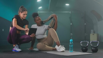 Fitness gym instructor showing workout plan on tablet to young African American woman in sportswear with towel. They are sitting on the gym floor, resting after workout.