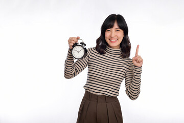 Smiling cheerful attractive young asian woman wearing sweater shirt holding alarm clock, point finger above and looking camera isolated on white background, studio portrait