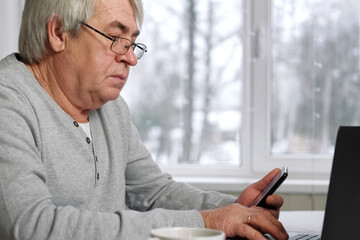 Handsome serious senior man wearing glasses using mobile phone while sitting at desk at his cozy workplace with laptop. Retired male working from home chatting with colleagues. Old Man with Technology