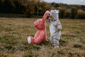 Little child wearing a plush mouse costume, playing with a big plush bunny toy, outdoors, in an...