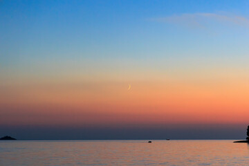 Fishing boats sail across the sea near Rovinj in Croatia at dusk while a narrow crescent of the waxing moon is in the sky