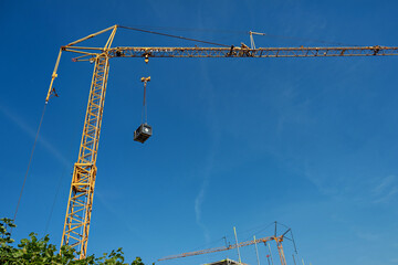 A tall yellow construction crane with a dangling black box against a blue sky in Germany.