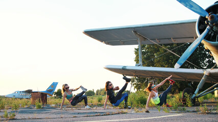 Beautiful, athletic, young women in sunglasses, in tights, perform synchronously different strength exercises, jumps, push-ups, lifting legs.on an abandoned airfield, near plane