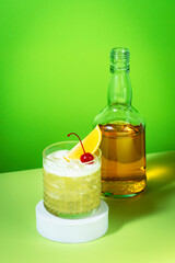 Whiskey sour cocktail with bourbon, lemon juice, egg white and ice, rocks glass and bottle on green background