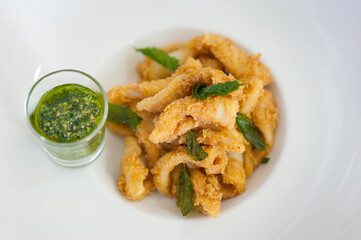 Close-up of calamari rings with fried rucola and green salsa brava. Battered squid appetizer. Seafood meal on a white plate. Horizontal food background. Restaurant and cafe menu concept.