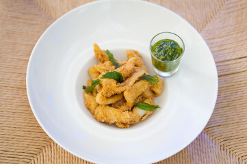 Close-up of calamari rings with fried rucola and green salsa brava on a white plate. Battered squid appetizer. Seafood meal on a rattan placemat. Horizontal food background. Restaurant menu concept.