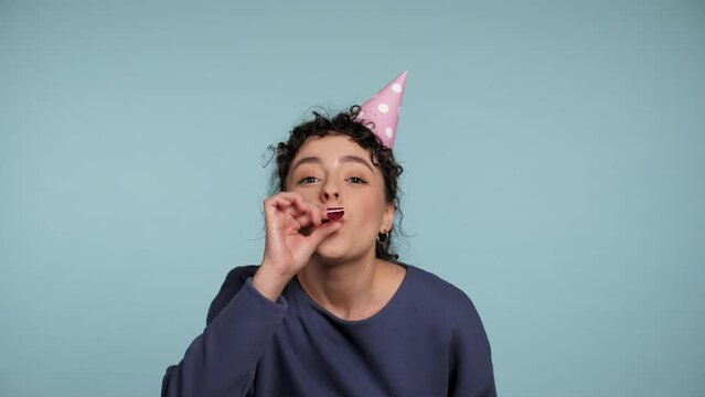 Close up happy curly woman in birthday cap blowing whistle celebrating holiday on isolated light blue background with copy space. Positive smiling cute female wearing blue sweater. People emotions