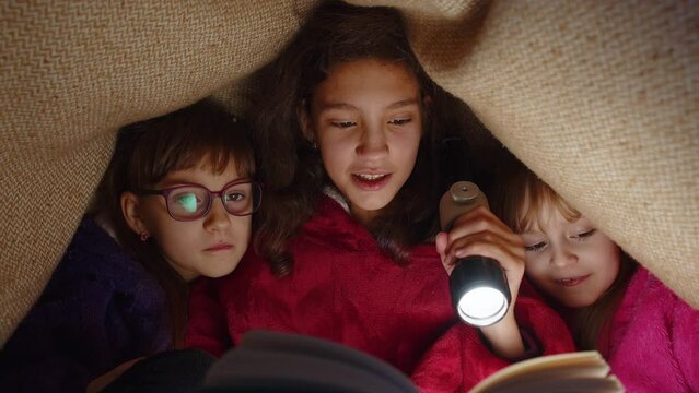 Teen girl and toddler child sisters under blanket plaid covers with flashlight reading interesting fairytale story book using flashlight. Female three siblings children kids friends in wigwam at home