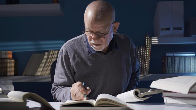 Wise old man reading a book, researching historian mature man.
Researcher or historian mature old man reading a book, taking notes while working in his study.
