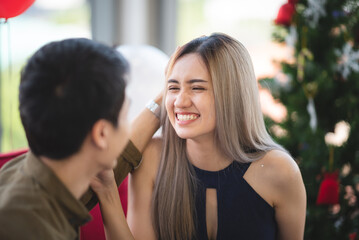 Cheerful and beautiful young Asian woman wearing New Year cap during Christmas Eve opening and receiving presents with handsome friend and boyfriend at home decorated with tree and balloons