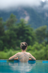 Rear view of young beautiful Asian woman in swimsuit relaxing while swimming in pool and leaning...