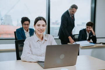 Focused and successful young female financer and marketing manager working and typing on laptop sitting in modern office with colleagues working in background