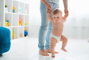 Cute little baby in a diaper learning to walk in a white room , mom holding his hands - obrazy, fototapety, plakaty