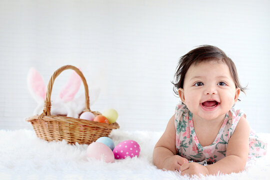 Portrait of six months crawling baby Easter egg basket with bunny ears on fluffy white rug, happy smiling adorable sweet little girl kid lying on white room, childhood baby Easter celebration concept.