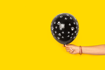Mano de mujer sosteniendo un globo negro con huellas de patas de gato y o perro sobre un fondo amarillo brillante liso y aislado. Vista de frente y de cerca. Copy space 