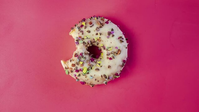 Eating delicious sweet donut with colorful sprinkles on pink background. Timelapse. Bakery and food concept. Top view of colorful frosted doughnut in 4K, UHD