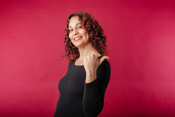 Young redhead woman wearing black ribbed dress isolated over red background pointing thumb finger back behind on workspace area copy space. Looking at the camera.