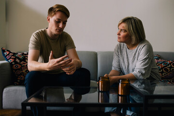 Mature parent looking at sad son sitting on sofa at home