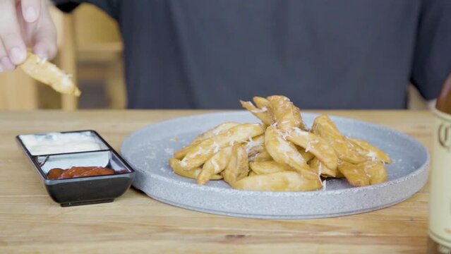 Plano a detalle de Plato de papas gajo con queso sobre una mesa de madera untando aderezo.