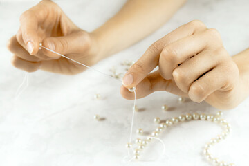 Close-up of woman hands stringing pearls on a necklace thread on white background. Beads jewellery...