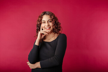 Fototapeta premium Young redhead woman isolated over red background enjoys talking touches face while laughing and looks at you tenderly. Laughing and smiling, showing genuine happiness and joy, positive emotions.