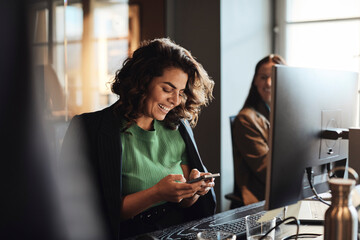Happy female entrepreneur using mobile phone sitting at desk in office