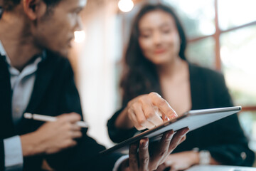 Smiling and laughing young Asian successful business woman discussing work with male colleague and partner in modern office while using digital tablet