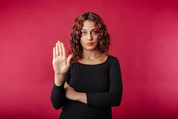 Portrait of young redhead woman wearing black ribbed dress isolated over red background doing stop...