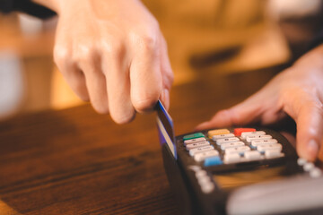 Closeup of hands of a young female cashier at barista swiping bank debit and credit card on NFC technology machine while processing payment for beverage and food ordered by customer