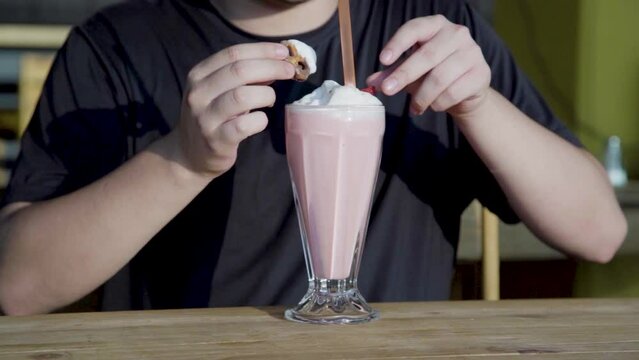 Hombre bebiendo una rica malteada de algod&oacute;n de az&uacute;car decorada con chocolate, pretzeles, cerezas y crema batida	en mesa de madera al atardecer.