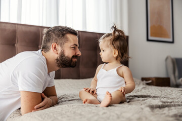 A serious baby girl is sitting on a bed and looking at her father while he is smiling at her.