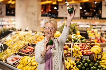 A happy senior woman is dancing with avocado in supermarket.