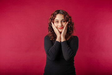 Fototapeta premium Young beautiful woman wearing black ribbed dress isolated over red background touch palms cheeks look you enjoy. Touching chin with palms, looks lovingly at the camera, at you.