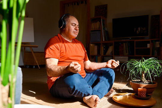Mature middle-aged overweight man in wireless headphones relaxing at home with guided meditation, listening to relaxing music and meditating in lotus pose.