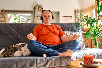 Overweight man in wireless headphones practicing yoga and guided meditation at home sitting in...